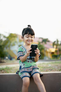 Girl using phone while sitting on retaining wall against sky