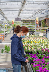 A young woman in mask chooses and buys seedlings in a garden center whith plants