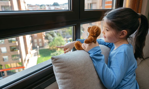 Girl showing the street to her teddy bear through the window