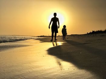 Silhouette father and daughter walking at beach against sky during sunset