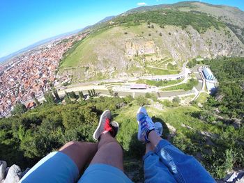 Low section of couple sitting on cliff by city and mountains