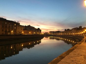 Illuminated buildings by river against sky at sunset