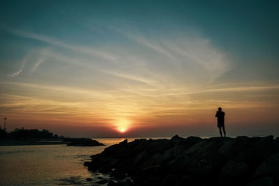 Silhouette man standing on rock by sea against sky during sunset