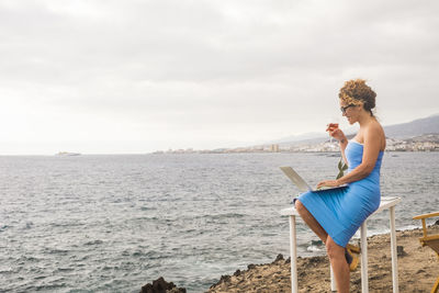 Woman using laptop while drinking wine at beach against sky