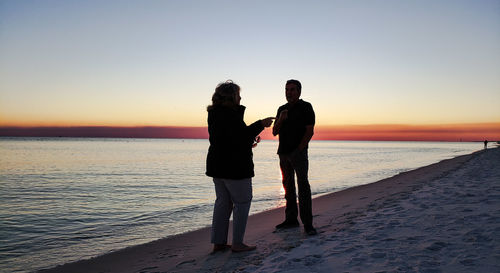 Silhouette friends talking while standing on beach against clear sky during sunset