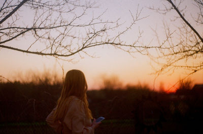 Woman standing by bare tree on field against sky during sunset