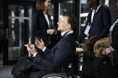 Side view of disabled businessman gesturing while sitting in wheelchair by delegates at seminar