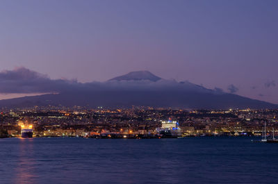 Illuminated city by sea against sky at dusk