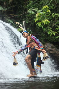 Side view of young man wearing traditional clothing standing against waterfall in forest