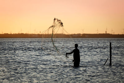 Silhouette fisherman throwing fishing net in sea against sky during sunset