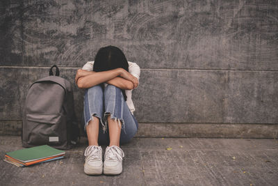 Depressed woman sitting by backpack and books against wall on footpath