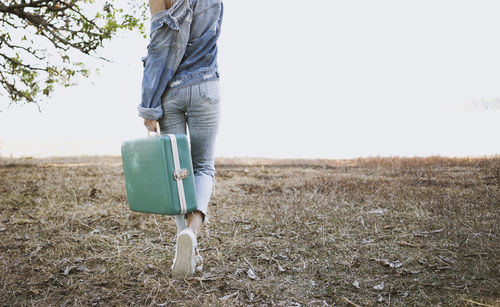 Low section of woman holding briefcase while walking on field against clear sky