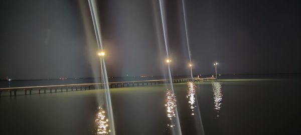 Illuminated street lights by river against sky at night