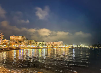 Illuminated buildings by sea against sky at night