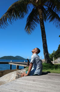 Man sitting on palm tree by plants against sky