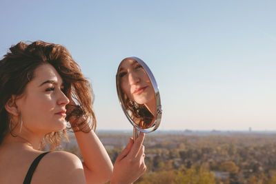 Side view of young woman looking in mirror against clear sky