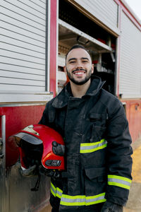 Cheerful bearded young firefighter standing near fire truck and holding red hardhat in hand while smiling and looking at camera