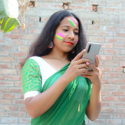 Beautiful young woman using phone while standing against brick wall