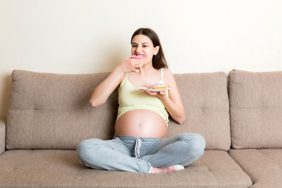 Smiling pregnant woman eating donut while sitting on sofa