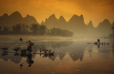 People with birds on boat in lake against mountains during sunset