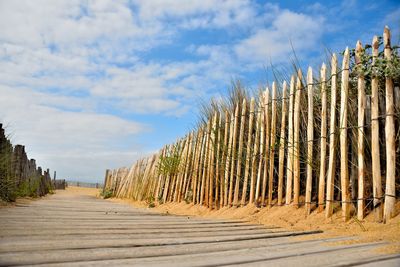 Wooden walkway leading towards beach against cloudy sky during sunny day