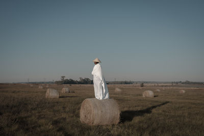 Young man covered with fabric standing on hay bale at agricultural landscape