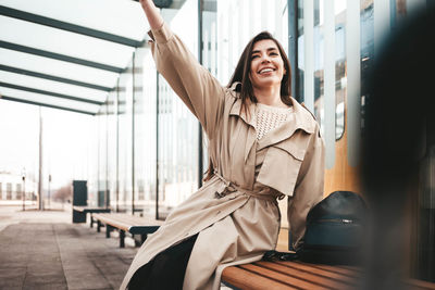 Positive young woman sitting at a public transport stop and waving