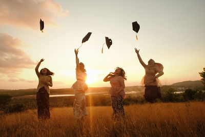 Silhouette people throwing mortarboards while standing on grassy field against sky during sunset