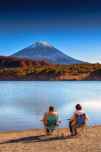 Rear view of men sitting on seat by lake against blue sky