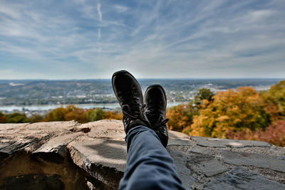 Low section of man relaxing on wall against cloudy sky