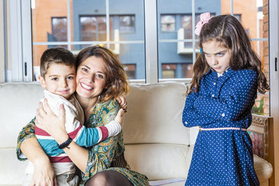 Portrait of smiling woman with son by angry daughter in living room at home