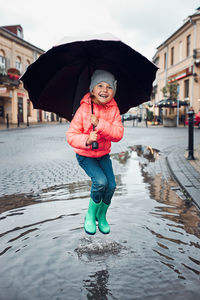 Happy smiling girl holding big umbrella jumping in the puddle during walk in a downtown