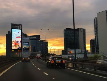 Cars on road by buildings against sky at sunset