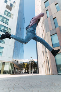 From below side view of young african american male in cool outfit jumping above ground on street while listening to songs in headphones and looking at camera