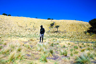 Full length of young man walking on grassy field against clear blue sky during sunny day