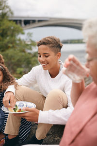 Smiling teenage boy having meal while sitting on lakeshore with family in park