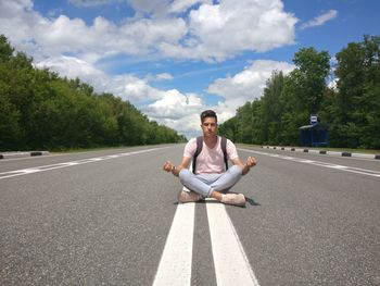 Full length of man with eyes closed meditating while sitting on country road against sky