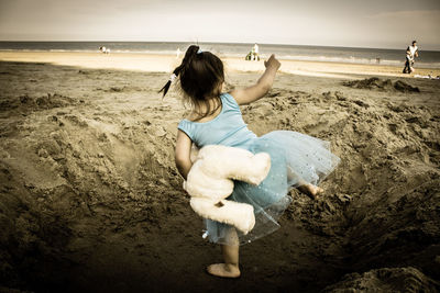 Full length rear view of girl holding stuffed toy while dancing at sandy beach