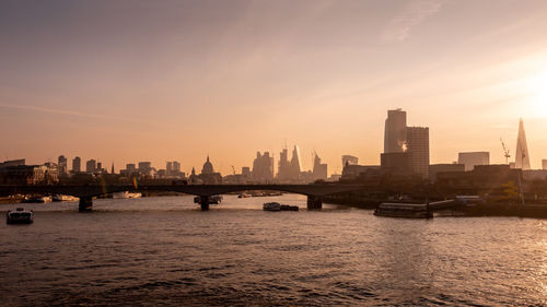 River by city buildings against sky during sunset