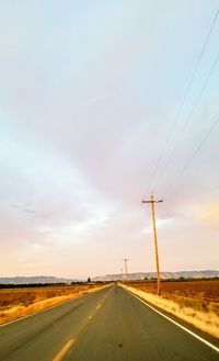 Road by electricity pylons against sky during sunset
