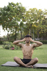 Full length of shirtless man sitting by tree