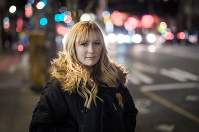 Portrait of young woman standing on sidewalk in illuminated city
