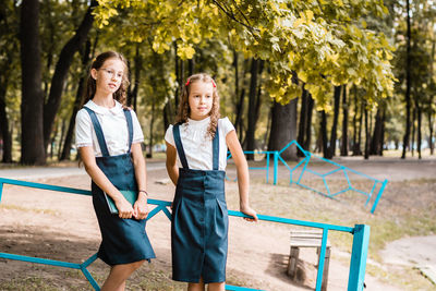 Two pupils in school uniform stand near the fence in the park on a warm day