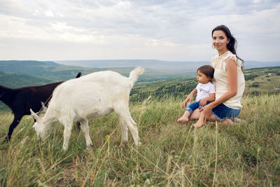 Woman with a child in a field on the mountain sitting next to a small goat in the summer