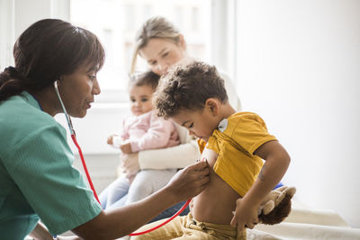 Side view of female doctor examining boy's heartbeat with stethoscope while mother sitting and daughter in clinic