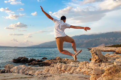 Full length of young man jumping on rock at sea shore against sky