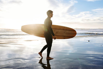 Side view of surfer man dressed in wetsuit walking on the beach with the surfboard in the morning with sunrise in the background