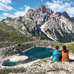 Rear view of couple sitting on rock by lake against sky