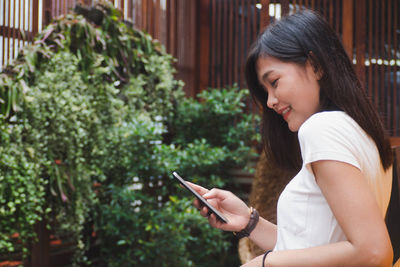 Side view of woman using phone while standing against plants