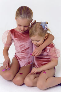 Two sisters sitting on a white background in pink bodysuits